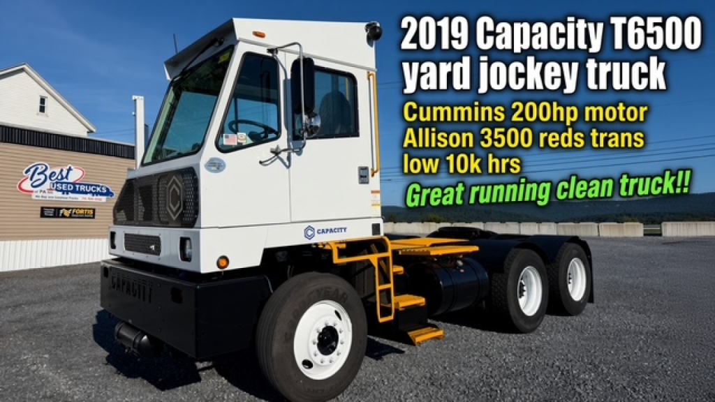 Truck side view showing 2019 Capacity TJ6500 yard jockey truck with white cab and yellow frame, Cummins 200hp motor text overlay.