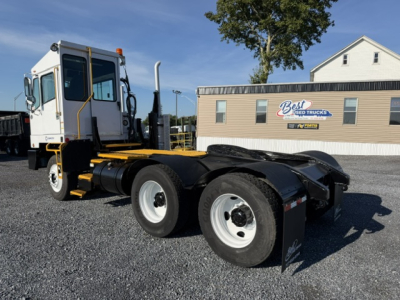 Truck chassis with attached cab and dual rear axles in yard, white cab with black accents, yellow frame, and storage area; sign in background reads Best Used Trucks.