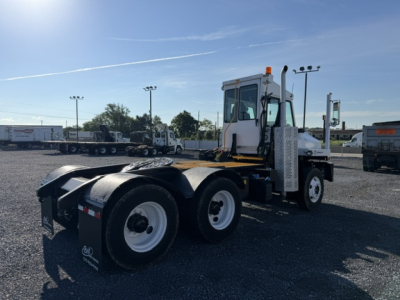 Truck tractor chassis with white cab and dual rear axles, visible text on flags and mirrors; DOT equipment with orange beacon, yard setting.