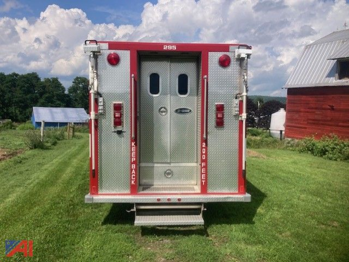 Rear view of a red and silver GMC 7000 rescue truck with double rear doors, '295' badge, and steps. Visible is a grassy field, a red barn, and a small blue tent in the distance.