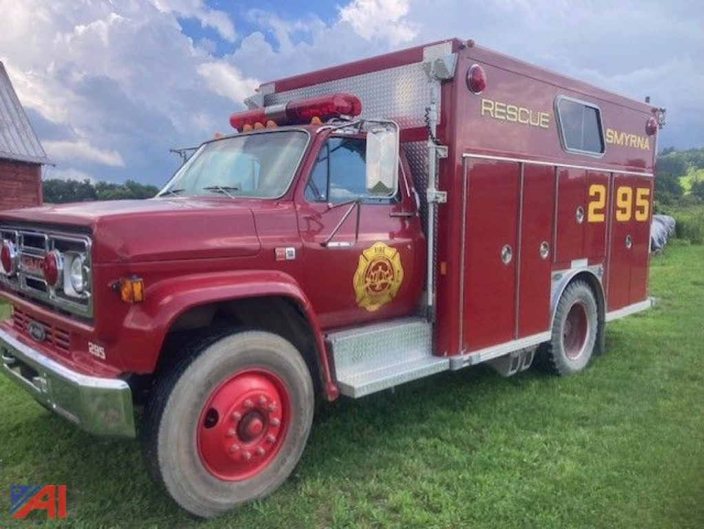 1987 GMC 7000 rescue truck side view showing red fire apparatus with gold rescue emblem and numbers 2 95 on rear quarter, EMS-style door insignia, silver step plates, and emergency lights.