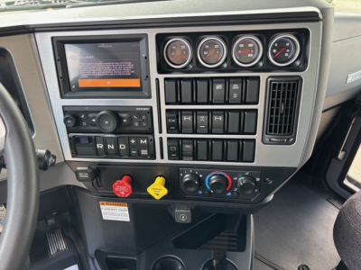 Dashboard of a Mack GR64F showing a touchscreen display, multiple gauges, climate controls, and switches. Visible text includes 'R N D + -' gear selector area and various warning labels.
