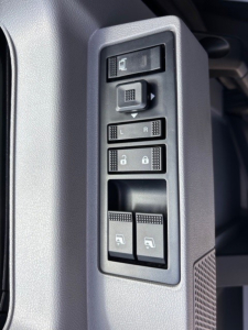Truck interior door panel showing window/lock controls and mirror adjust buttons on a 2026 Mack GR64F cabin. Includes L/R mirror selection, window switches, and lock buttons.