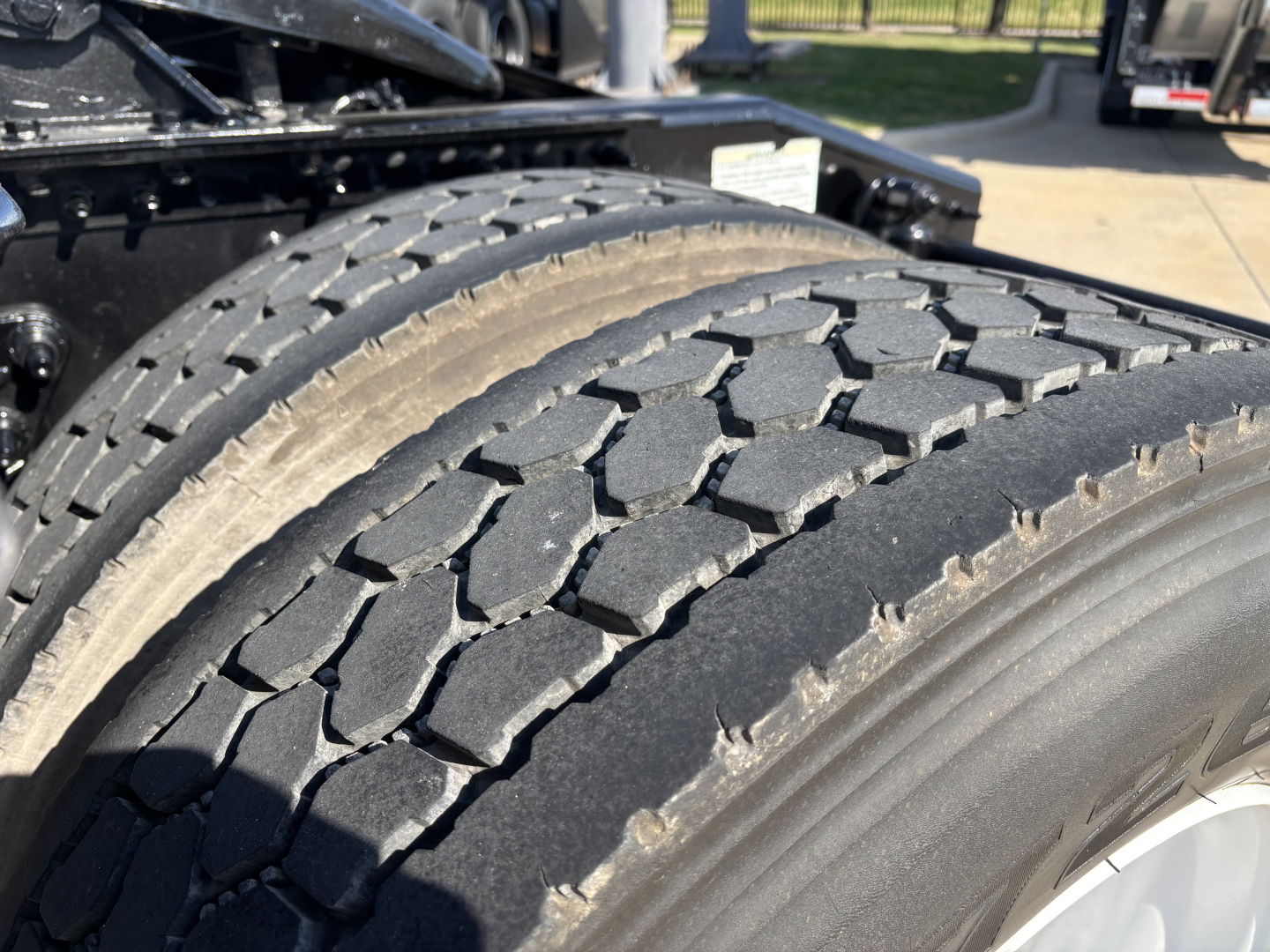 Truck tire close-up showing tread pattern on dual rear axle with black rubber and light reflecting on sidewall; hints of chassis in background.
