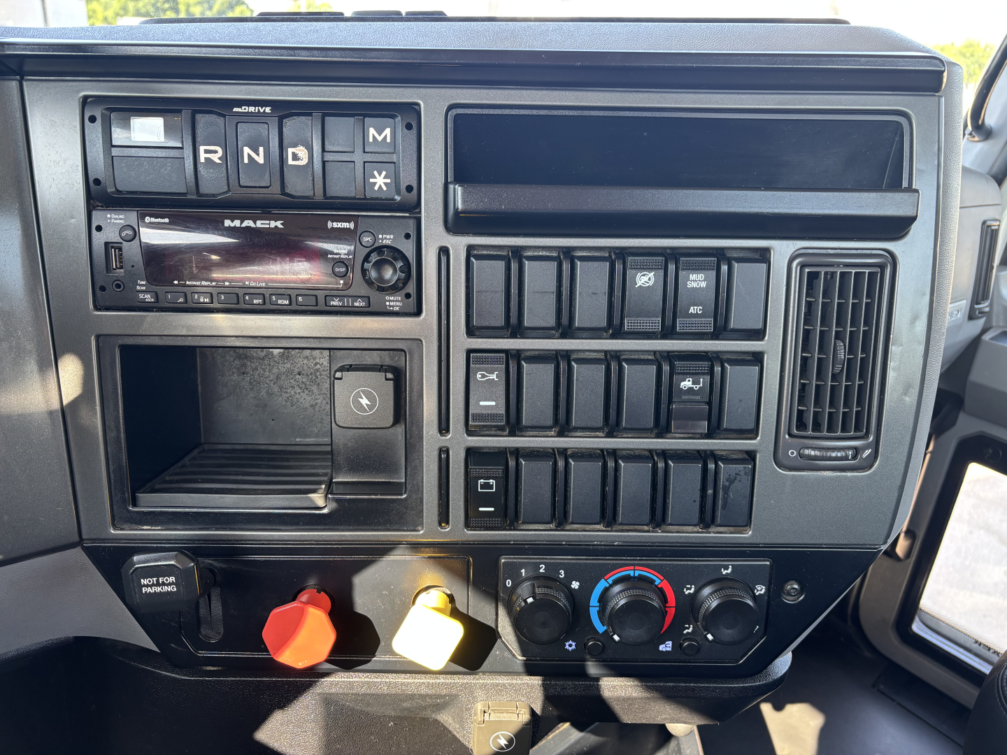 Dashboard panel of a Mack AN42T truck showing gear selector R N D M, radio with Mack branding, multiple switches, climate controls, and two colored knobs (orange and yellow) at bottom left.