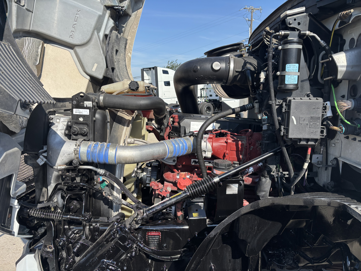 Truck engine bay of a 2022 Mack AN42T showing red engine, blue hoses, black tubing, electrical wiring, and fluid reservoirs. Various components and a metal frame are visible.