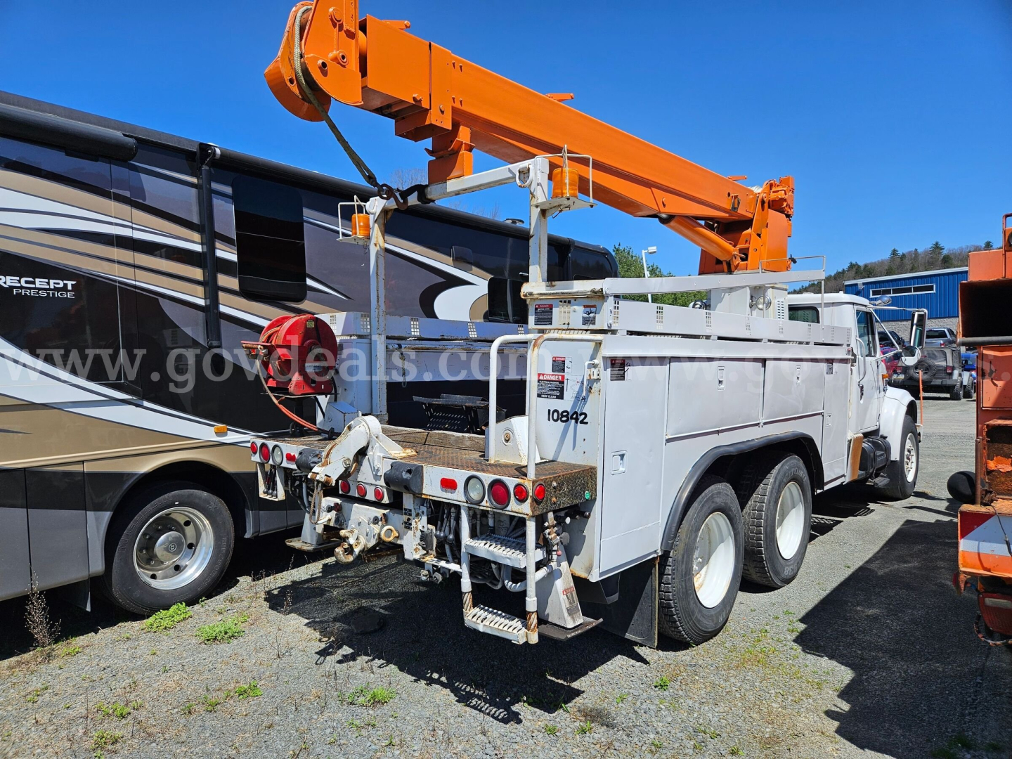 Back view of a white International 4900 boom truck with an orange hydraulic crane, mounted tools, and dual rear axles in a lot.