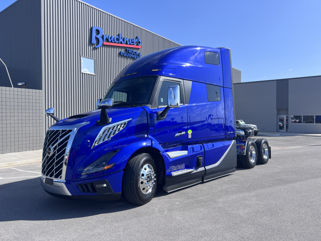 Blue Volvo VNL64T860 tractor-trailer tractor in front of a dealership building with 'Bruckner Truck Center' sign. Large chrome grille, aerodynamic cab, side mirrors, and dual rear axles visible.