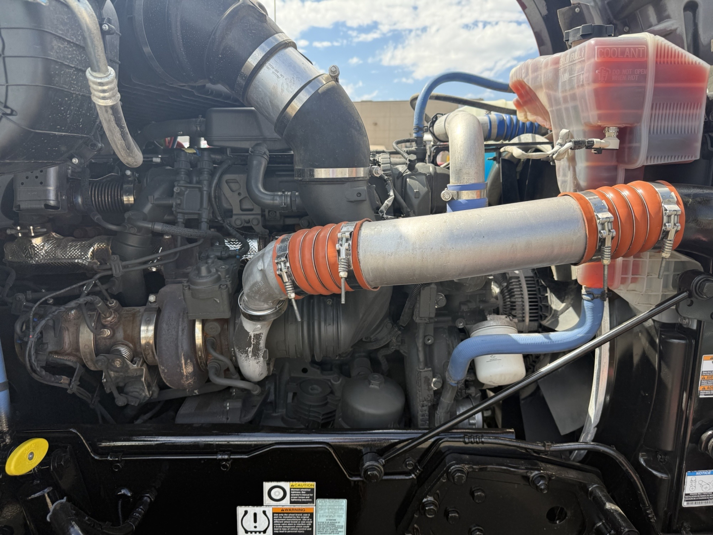 Kenworth T880 engine bay showing large turbocharger piping, orange silicone hoses, silver intake pipe, blue coolant line, translucent red coolant reservoir, and various engine components.