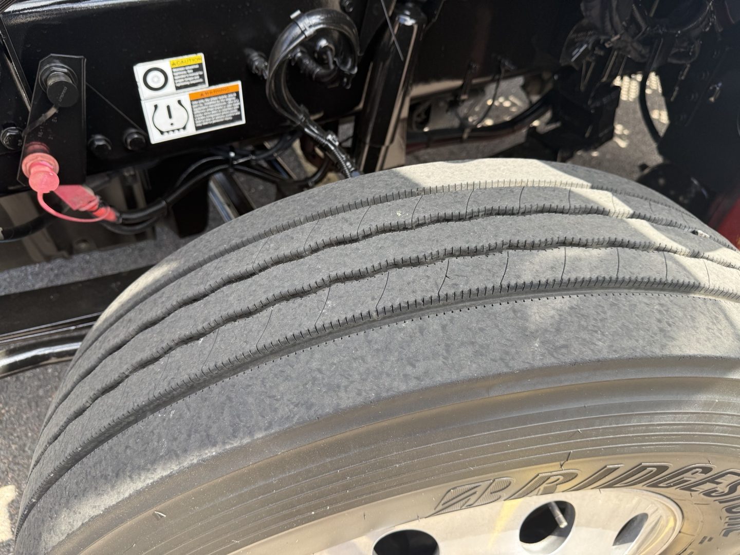 Kenworth T880 wheel and front suspension area showing dual rear tires, tire tread, and part of chassis with warning labels and wiring.