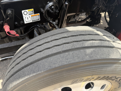 Kenworth T880 wheel and front suspension area showing dual rear tires, tire tread, and part of chassis with warning labels and wiring.