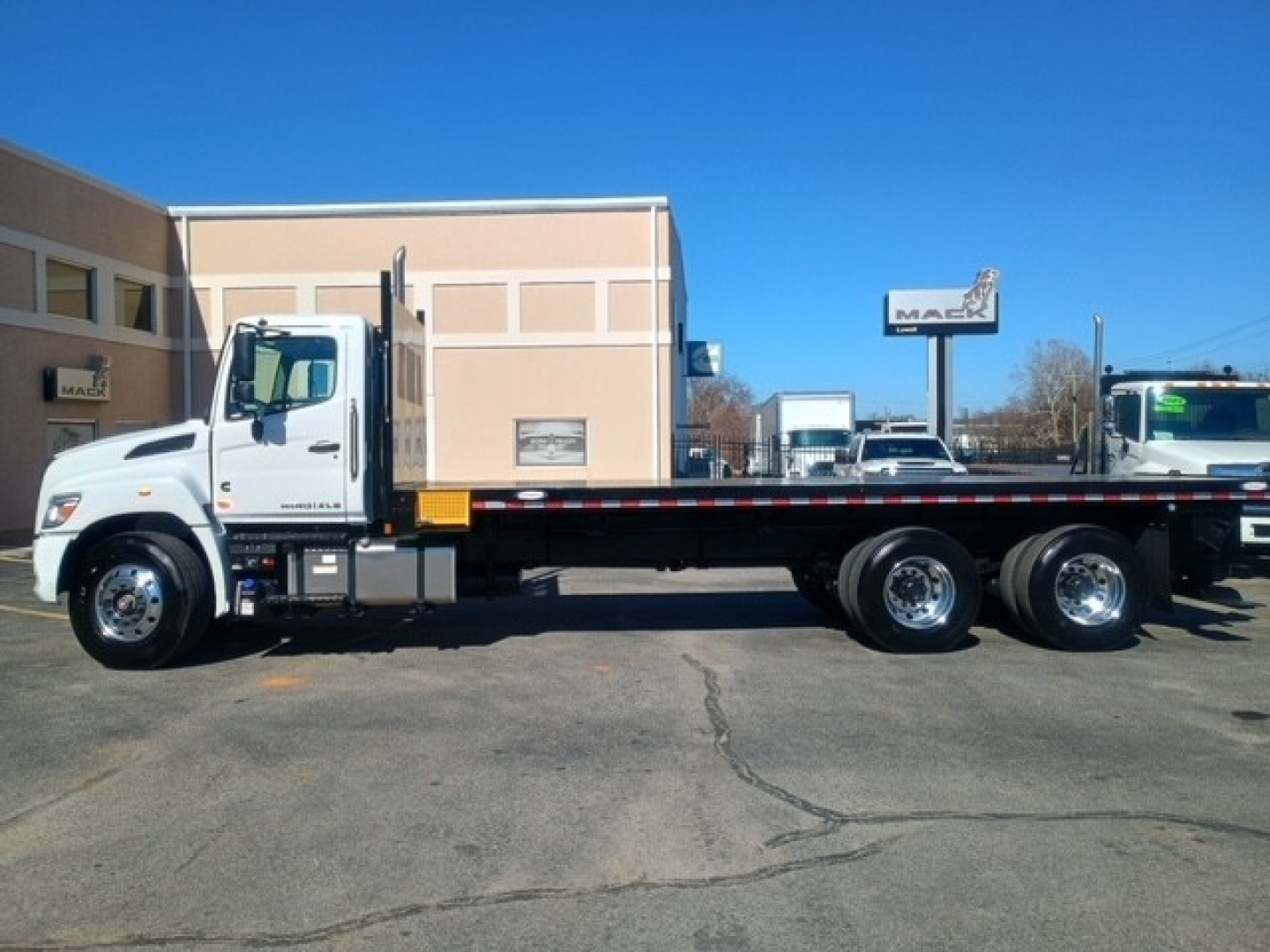 Flatbed truck side view showing cab with white paint, long heavy-duty flatbed, dual rear wheels, and chrome wheels. No visible branding text legible in image.