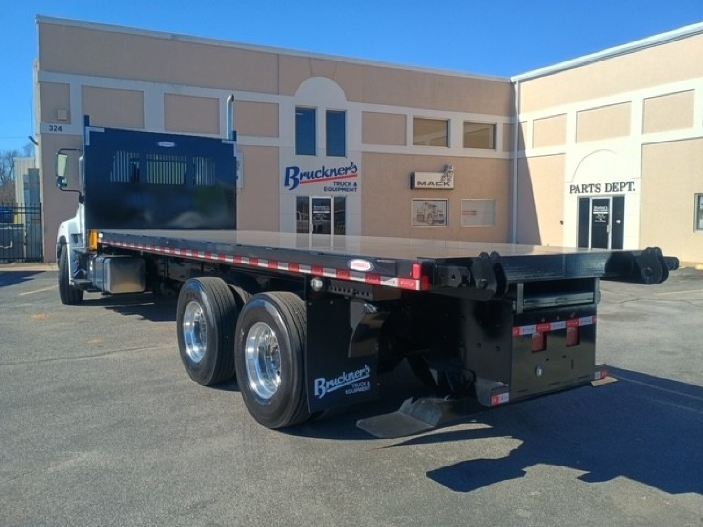 Flatbed truck trailer with dual rear axles and blue tool/controller box in yard; background shows office building and parts dept.