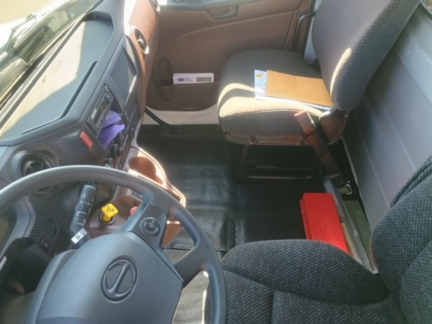 Hino XL8M interior view showing driver seat, steering wheel, dashboard, and passenger seat with documents and a red tool case.