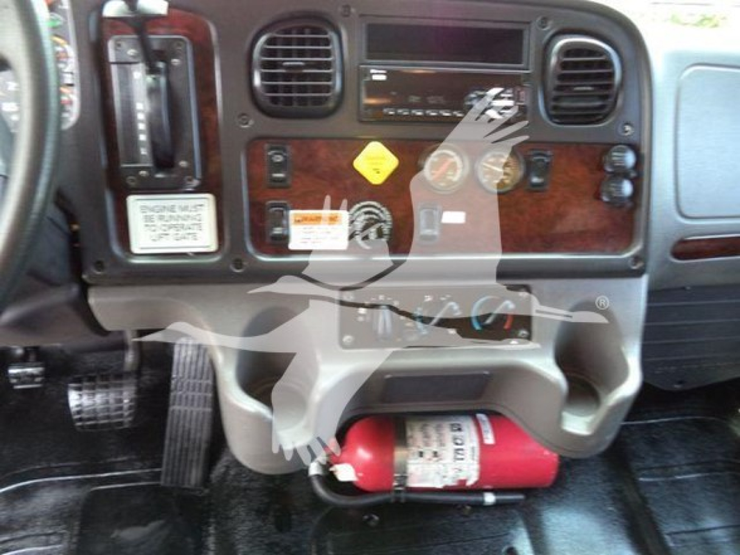 Dashboard of a Freightliner M2 106 cab showing wood grain panel, steering column, various gauges, warning stickers, and a red fire extinguisher mounted under the dash.