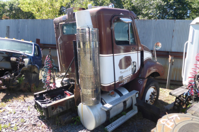 Truck cab rear-side view showing brown and white Freightliner CL112 daycab with chrome cylindrical exhaust, fuel tank, and ladder steps.