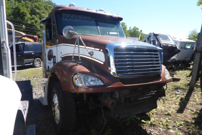 Truck front view showing Freightliner CL112 cab with brown and white paint, chrome grille, and large windshield in a salvage yard.