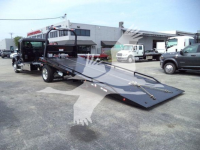 Kenworth T-Series rollback tow truck with flatbed and ramps, outdoor lot, front view showing raised bed and wheel chocks.