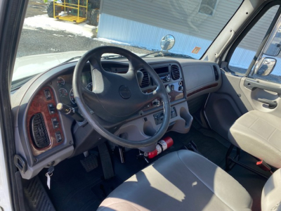 Interior of a Freightliner M2 106 cab showing steering wheel, dashboard, and driver seat with a fire extinguisher mounted under the dash.