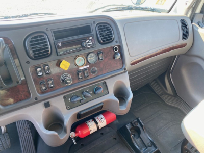 Cab interior of a Freightliner M2 106 dash with wood-grain paneling, gauges, knobs, a red fire extinguisher under the dashboard, and cup holders.