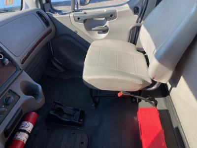 Cab interior of a Freightliner M2 106 truck showing a light gray fabric driver's seat, dashboard with glove compartment, and red tool/kit box on the floor near the seat.