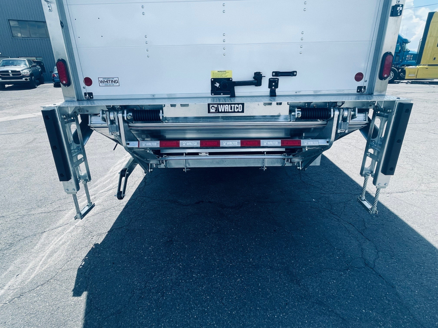 Rear view of a new Mack MD6 box truck showing tailgate, ramps, and undercarriage components.