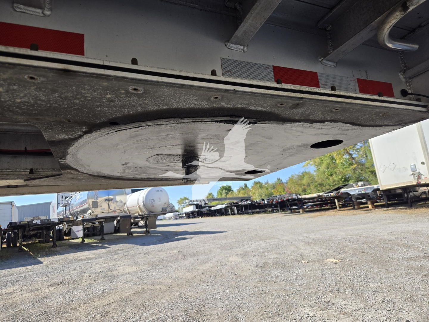 Trailer underside showing a circular drain/plate and welded framework with reflective tape on edge; yard with other trailers in background.