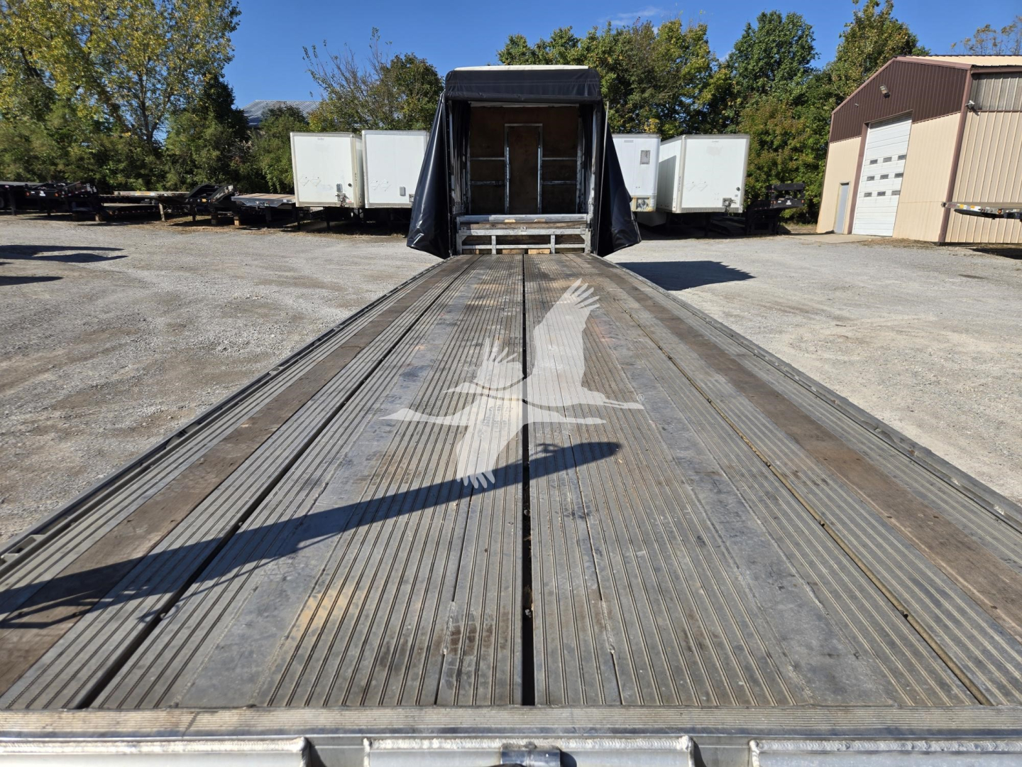 Flatbed trailer in-service showing long metal deck with dual grooves and ramp structure at rear, observation of trailer bed and surrounding yard.