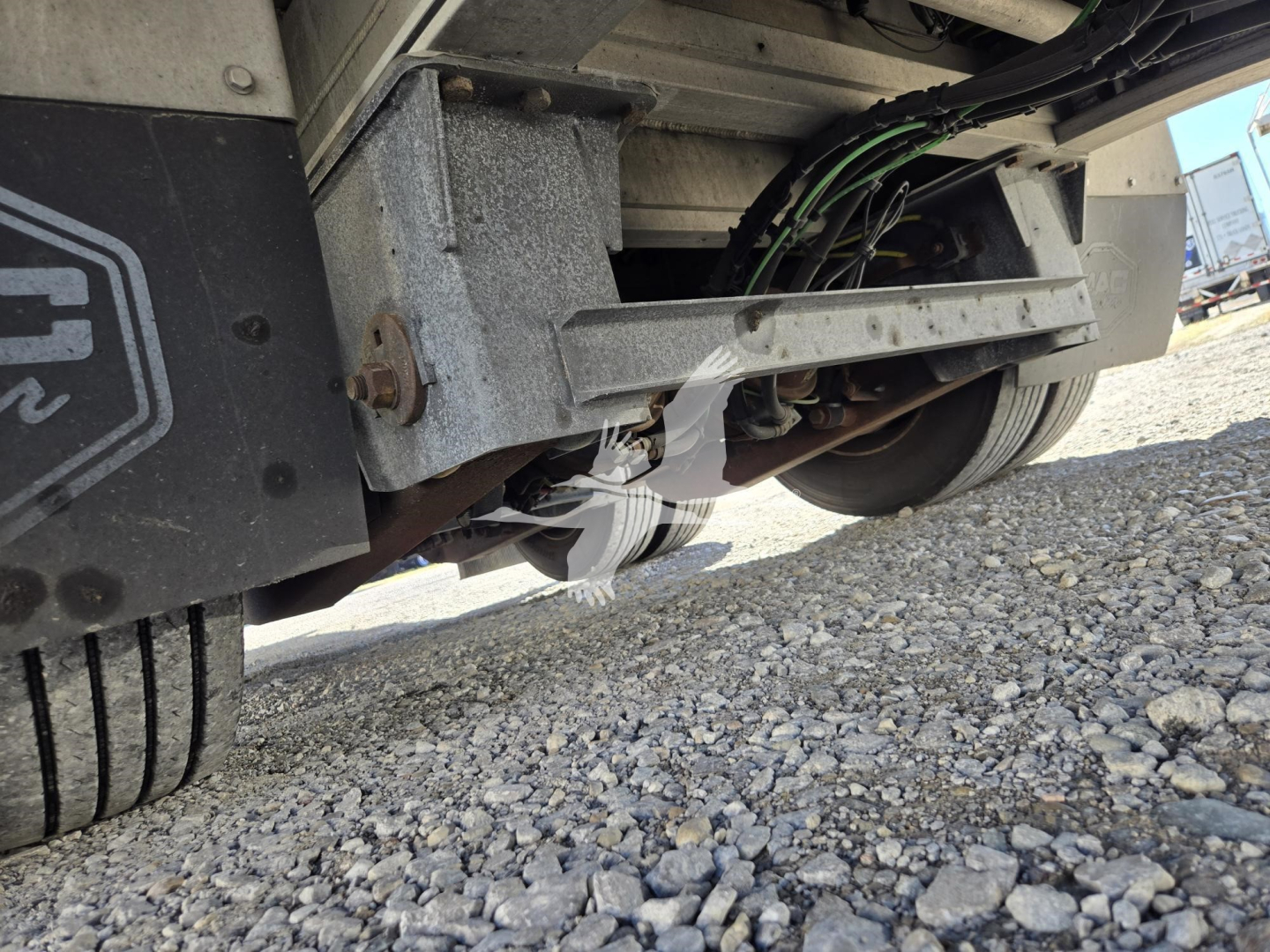 Long view of a 2019 Mac drop deck trailer undercarriage showing wheel, suspension, and hydraulic lines with gravel ground.
