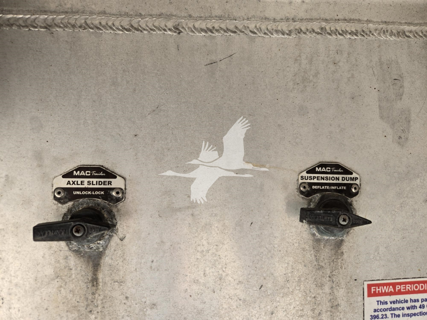 Mac trailer deck showing axle slider and suspension dump levers with a white bird decal in center.