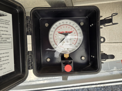 Envelope-style dial gauge in a black protective case mounted on a trailer hitch with a red button and small label, showing a circular Right Weigh scale inside the lid.
