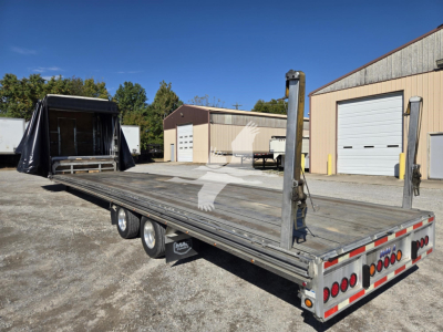 Flatbed trailer with a drop deck, three axles, and a ramp at back; shows metal deck, rear lights, and a vertical loading mechanism near the front.