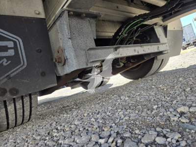 Long view of a 2019 Mac drop deck trailer undercarriage showing wheel, suspension, and hydraulic lines with gravel ground.