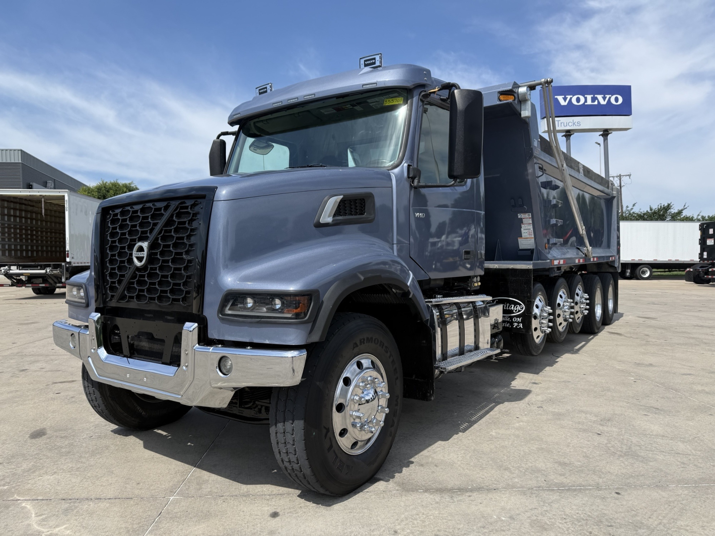 Volvo dump truck with 6x4 dual rear axles and chrome bumper, front grille emblem, in-service exterior, blue-gray color, Volvo dealership in background.