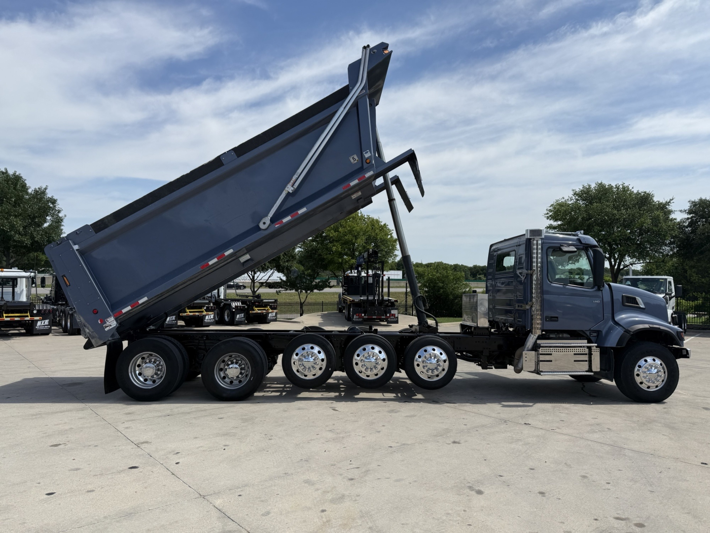 Volvo VHD dump truck side view showing dump bed raised, multiple rear axles, blue truck with chrome wheels.