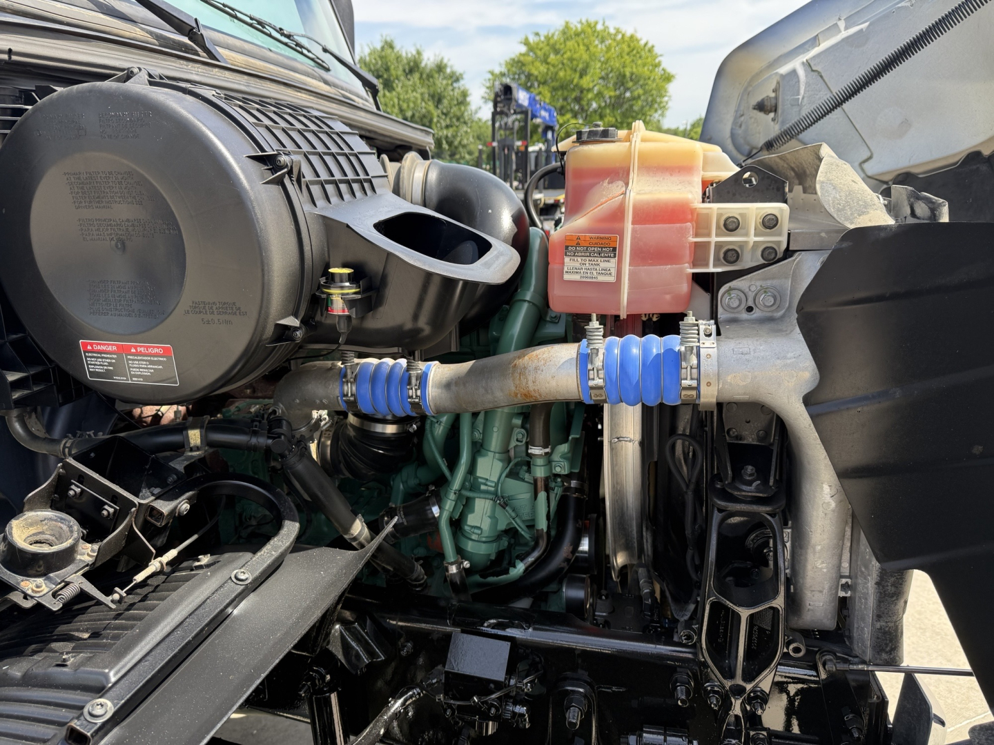 Volvo VHD powertrain area showing engine, radiator reservoir, hoses, and blue silicone hose with clamp in engine bay.