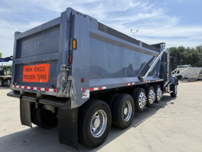 Truck rear three-quarter view of a blue Volvo dump truck with multiple axles and a tailgate, showing fuel and warning placards and rear lights.