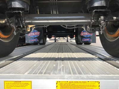Flatbed trailer undercarriage showing axle beams, tires, and Utility-brand mud flaps with 1200 sticker, in a truck transport bed.