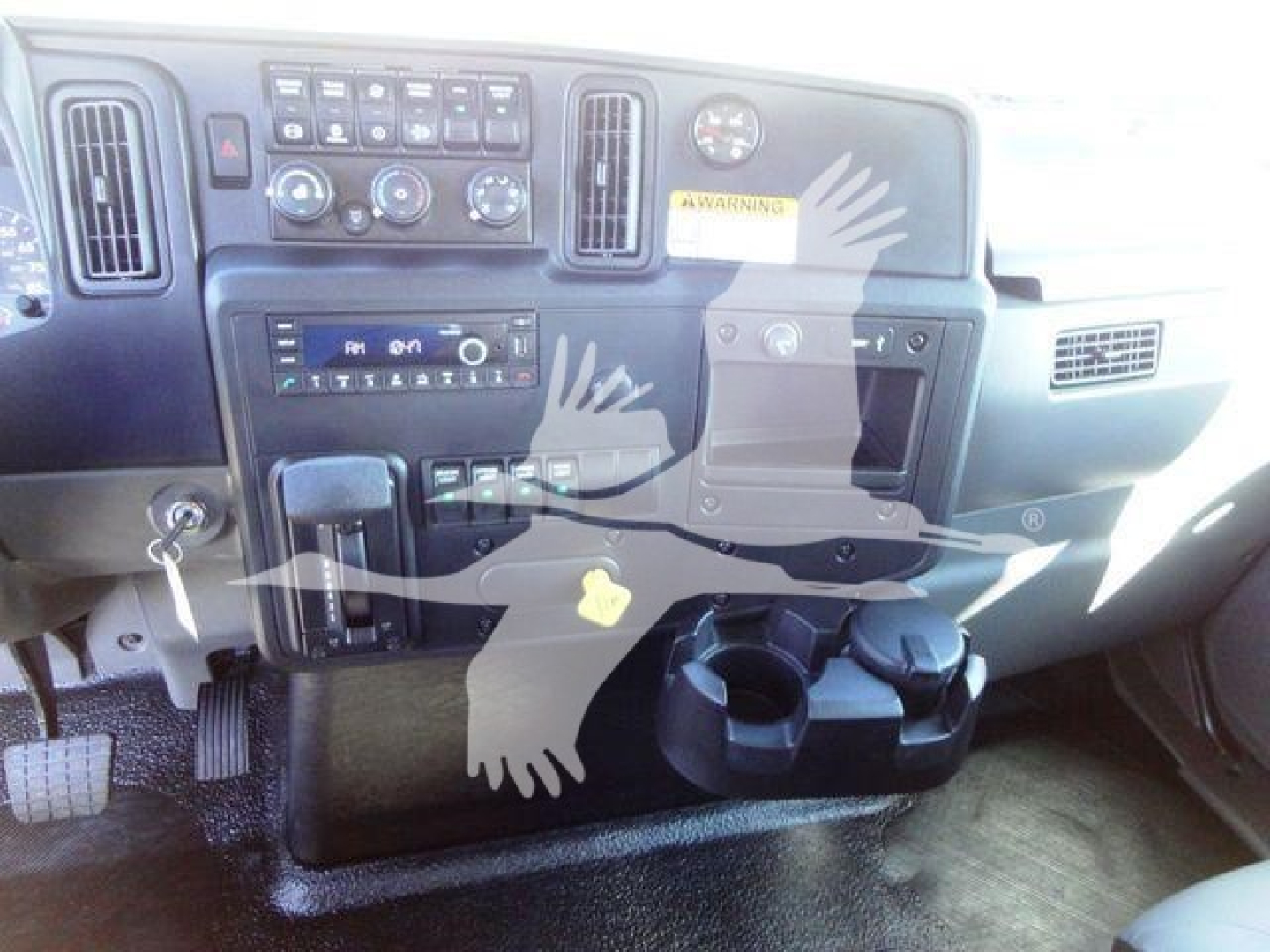 Truck cab dashboard with controls, vents, cup holders and an ignition key inserted; dashboard features climate controls, radio, and warning sticker.