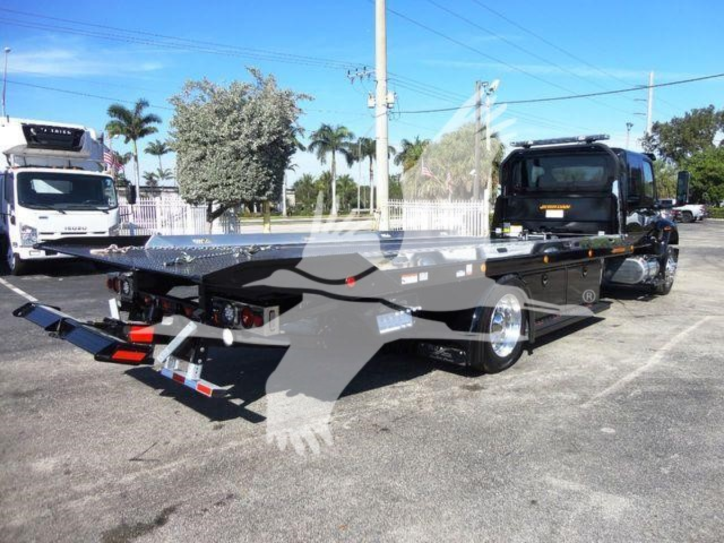 Tow truck rollback bed with black chassis and silver accents, rear view showing goose-neck hitch and lighting; dealership lot in background.