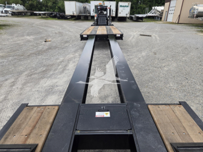 Lowboy trailer deck with long black I-beam frame and wooden deck sections; Fontaine 60Hd MX trailer in distance; VIN 13NE58306V35A7290 stock V35A7290.