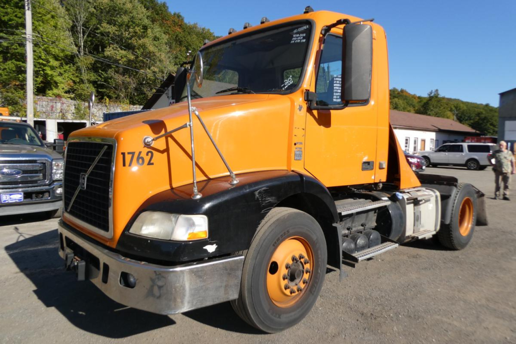Orange Volvo VNM cab forward truck with black lower front, showing '1762' on fender and Volvo emblem; side mirrors; front bumper; daytime lot with other trucks.