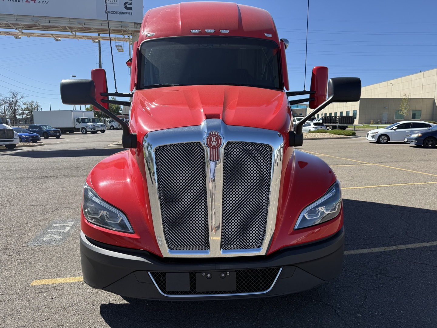 Kenworth T680 truck front view showing chrome grille, red hood, black bumper, and side mirrors in a parking lot.