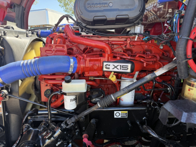 Kenworth T680 engine bay with bright red engine block and X15 badge, blue air intake hose, white coolant reservoir, and Pacar filter housing visible.