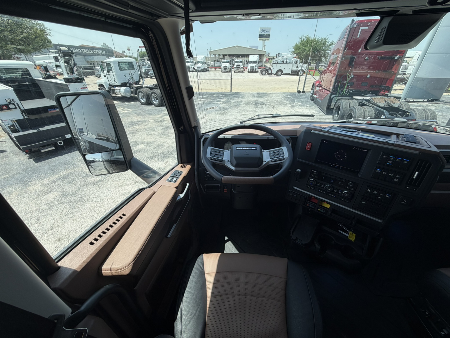 Cabin view inside a new 2027 Mack PR64T76H truck showing steering wheel, dashboard, center console, and brown seat upholstery.