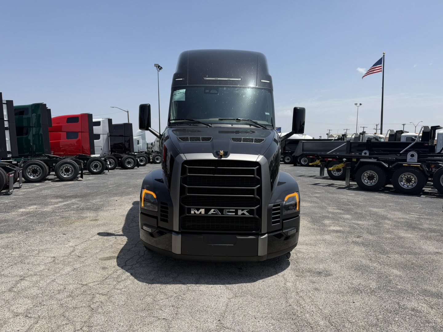 Mack PR64T76H truck front view showing bold grille with Mack emblem, large bumper, and headlight clusters; surrounding lot with other trucks.