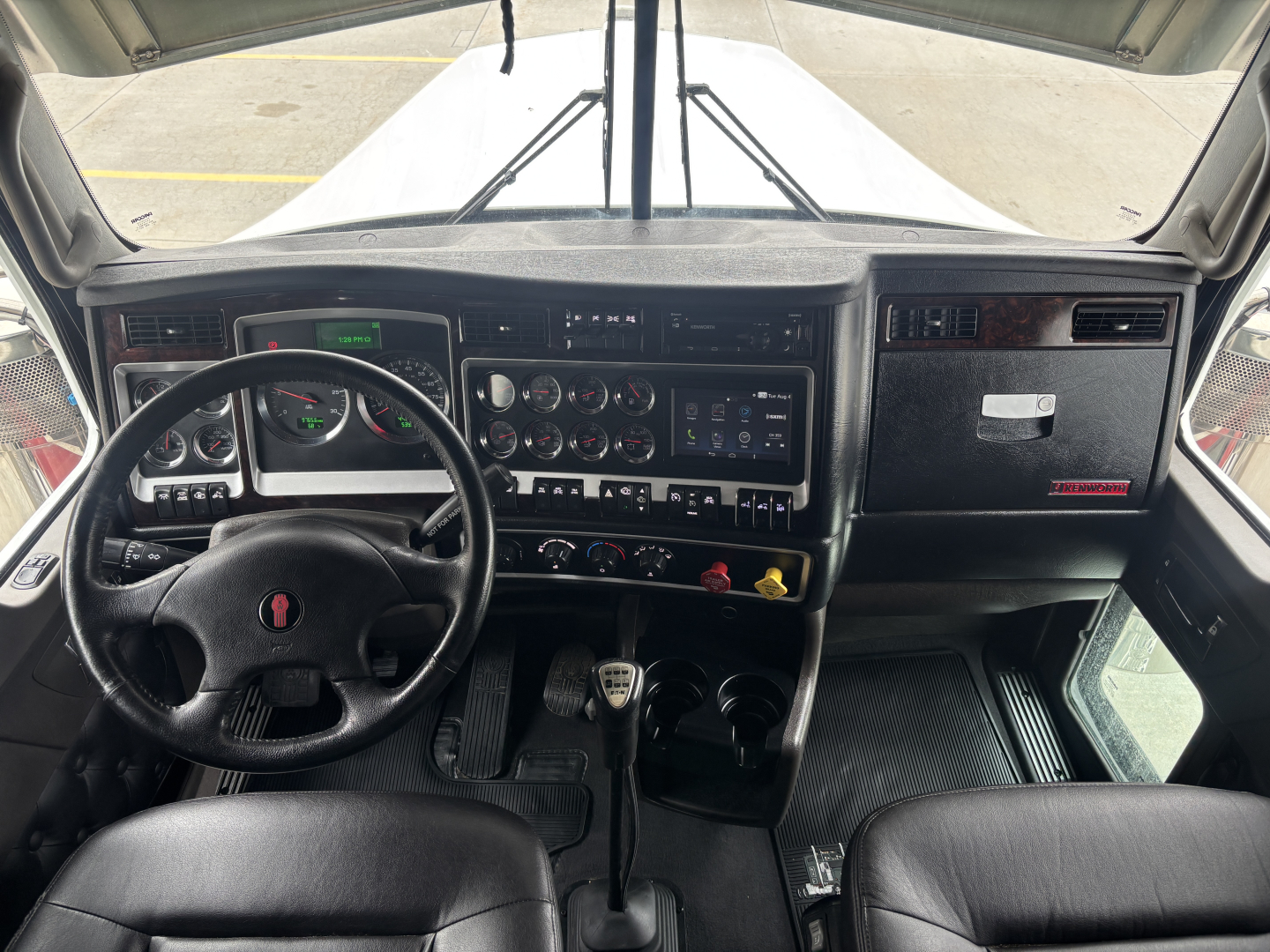 Kenworth W900L cab interior showing dashboard with gauges, steering wheel, and center control panel; manual shifter and dash accessories visible.