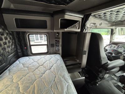 Kenworth W900L cab interior showing sleeper area with quilted padding, overhead compartments, and driver seat; dashboard visible at right.