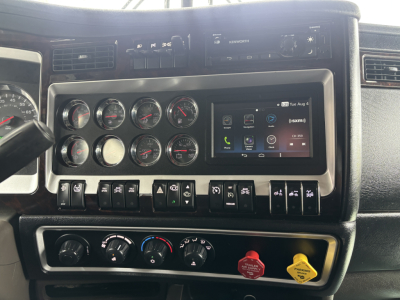 Dashboard panel of a Kenworth W900L showing multiple gauges, a touchscreen infotainment unit, control switches, and climate controls.