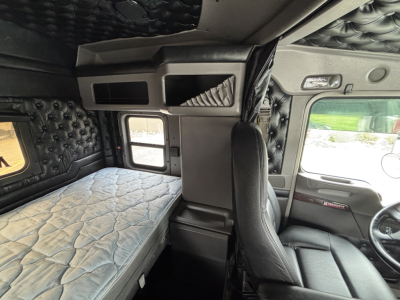 Kenworth W900L sleeper cab interior showing bunk bed and captain chair with diamond-stitched panels and overhead storage compartments.